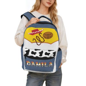 Puede incluir: Una mochila con textura de mezclilla azul que presenta un panel superior amarillo con un sombrero de vaquero rojo y una insignia marrón, una sección central blanca y un bolsillo con estampado de vaca en blanco y negro. El nombre CAMILA está impreso.