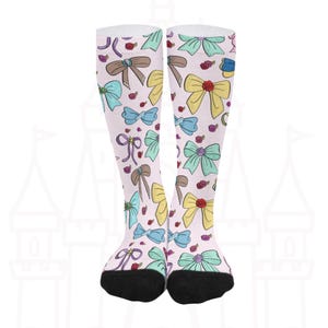 Könnte beinhalten: Kniestrümpfe mit hellrosa Hintergrund. Die Socken sind mit bunten Schleifen in Gelb, Blau, Grün und Braun sowie kleinen roten Rosen gemustert. Die Zehen sind schwarz.