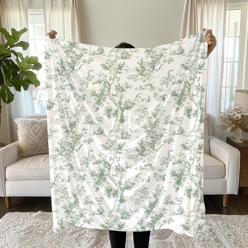 Op de afbeelding: Een witte deken met een groen toile de Jouy-patroon met bomen, dieren en figuren. De deken wordt omhoog gehouden, waarbij het volledige ontwerp wordt getoond. Het patroon herhaalt zich over de stof, waardoor een samenhangend en decoratief uiterlijk ontstaat.