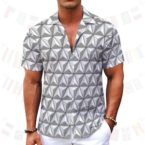 Puede incluir: Camisa de manga corta con un patrón geométrico 3D en tonos grises y blancos. La camisa tiene un cuello clásico y se combina con pantalones blancos. El estilo general es casual y moderno.