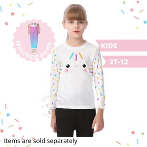 May include: A white long-sleeved shirt featuring a cartoon cat face and colourful sprinkles. The shirt is worn with black trousers. The image also shows matching trousers and text that says "KIDS 2T-12". The words "Items are sold separately" are also visible.