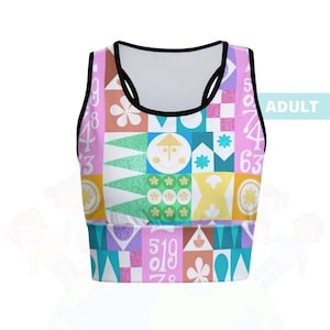 May include: A colorful crop top with a geometric pattern featuring numbers, flowers, and a smiling house design. The top has a black trim and the word "ADULT" is printed on the side. The design incorporates various colors including pink, blue, green, and yellow.