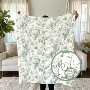Op de afbeelding: Witte deken met een groen toile de jouy patroon met Winnie the Pooh-personages en bossc&egrave;nes. De deken wordt omhoog gehouden en toont het volledige ontwerp. Een cirkelvormige afbeelding van Winnie the Pooh met een honingpot staat in de hoek.