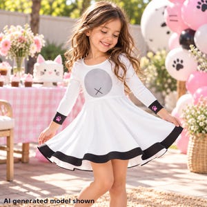 May include: A white long-sleeved dress featuring a round neckline, a grey circular graphic on the chest, black cuffs with small pink paw prints, and a wide black hemline. The skirt has a flared silhouette that falls above the knee.