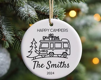 Op de afbeelding: Ronde witte keramische kerstversiering met een zwarte illustratie van een camper, een kerstboom en de tekst "Happy Campers" en "The Smiths 2024". De versiering hangt aan een beige touwtje.