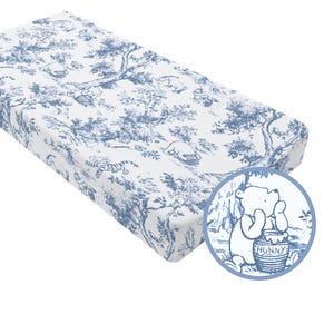 May include: A white and blue changing pad with a toile pattern featuring Winnie the Pooh characters and forest scenes. The pad has a rounded corner and a circular graphic of Winnie the Pooh with a honey pot.