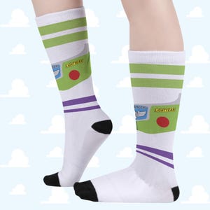 Puede incluir: Un par de calcetines blancos de caña alta con un estampado gráfico inspirado en un traje de ranger espacial. El diseño incluye detalles en verde lima, morado y negro, con un círculo rojo y el texto "LIGHTYEAR" impreso en el lateral.
