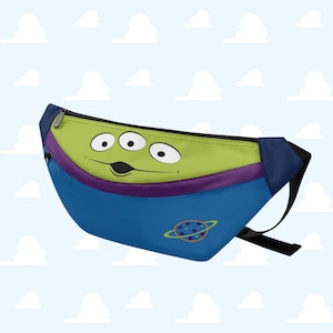 Op de afbeelding: Een blauwe heuptas met het gezicht van de Little Green Men uit Toy Story. De tas heeft een groene bovenkant met drie ogen, een paarse streep en een planeetgrafiek. Hij heeft een zwarte rits en riem.