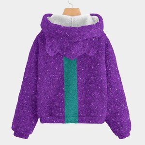 May include: A purple hooded sweatshirt with a glittery texture and a teal stripe down the center. The hood has a white, fluffy lining and small, rounded ear-like details. The sweatshirt is hanging on a wooden hanger.