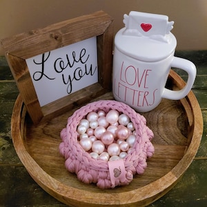May include: A wooden framed sign with the words "Love you" in black script sits next to a white mug with "Love Letters" in red lettering. A pink crocheted basket filled with pearls is on a wooden tray.