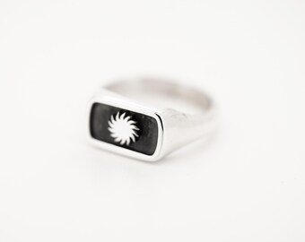 May include: A silver signet ring featuring a rectangular face with a black inlay. Centered on the black inlay is a small, raised silver sun emblem with radiating rays. The band of the ring is smooth and polished, maintaining a consistent metallic finish.