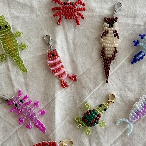 May include: A collection of nine animal-shaped charms crafted from small colorful beads. The charms include a red crab, a green lizard, a blue and black fish, a pink shrimp, a brown otter, a blue axolotl, a pink axolotl, a green turtle, and a fish.