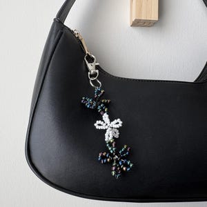 May include: A beaded bag charm featuring three flower-shaped motifs attached to a silver-toned metal lobster clasp. The top and bottom flowers are crafted from iridescent dark blue and purple beads, while the center flower is made of white beads.