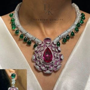 May include: A silver necklace with a large pink and white flower pendant and green teardrop-shaped stones. Matching earrings with a pink and white flower design and a green stone are also pictured. The necklace has the text "PREMIUM QUALITY" on it.