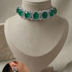 May include: An opulent emerald and diamond necklace and earring set displayed on a mannequin. The necklace features large, square-cut emeralds with a diamond halo. Matching earrings are also shown.