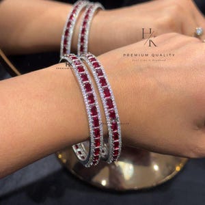 Può includere: Bracciali d'argento con una fila di pietre preziose rosse rettangolari e un bordo di piccole pietre bianche. I bracciali sono mostrati su un polso. Il testo "HK PREMIUM QUALITY Feel like a Diamond" è visibile.
