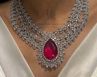 May include: A silver necklace with a large, teardrop-shaped ruby pendant surrounded by smaller diamonds. The necklace is made of multiple strands of diamonds and has a delicate, floral design.