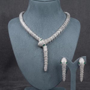 Puede incluir: Un conjunto de collar y pendientes de serpiente plateados. Las joyas presentan un diseño de serpiente con ojos de color esmeralda. El collar está colocado sobre un maniquí y los pendientes se exhiben en un soporte.