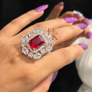 May include: An elaborate ring with a central, deep red gemstone, encircled by smaller, clear stones. The ring has a silver-toned setting and is shown on a hand with purple glitter nail polish. The ring has the text "HK PREMIUM QUALITY Diamond".