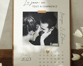 May include: A framed vertical poster featuring a black and white photograph of a couple, the text Le jour où TOUT A COMMENCÉ, the names Morgan & Elena, and a March 2023 calendar with a gold heart marking a specific date on the calendar grid.