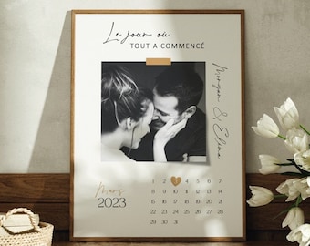 May include: A framed print with a black and white photo of a couple, a calendar for March 2023, and the text "Le jour où tout a commencé". The print is on a wooden dresser with a basket and a vase of white tulips.