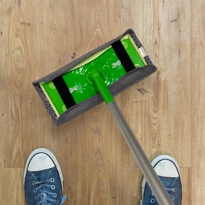 May include: A person wearing blue trainers is using a Swiffer WetJet mop to clean a wooden floor. The mop has a green cleaning pad and a grey handle.