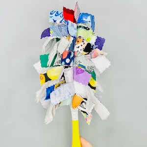 May include: A colourful fabric dust mop with a yellow handle. The mop is made of various scraps of fabric in different patterns and colours, including blue, green, red, and white.