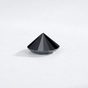 May include: A faceted, black gemstone with a conical shape. The gem has a flat base and a pointed top, with multiple facets creating a reflective surface. The gemstone is set against a white background.