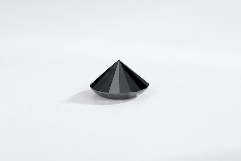 May include: A faceted, black gemstone with a conical shape. The gem has a flat base and a pointed top, with multiple facets creating a reflective surface. The gemstone is set against a white background.