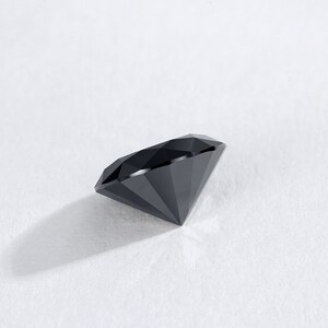 May include: A faceted, black gemstone with a brilliant cut. The diamond-shaped gem is displayed on a white surface, showcasing its sharp angles and reflective surfaces. The gemstone's dark color contrasts with the bright background.