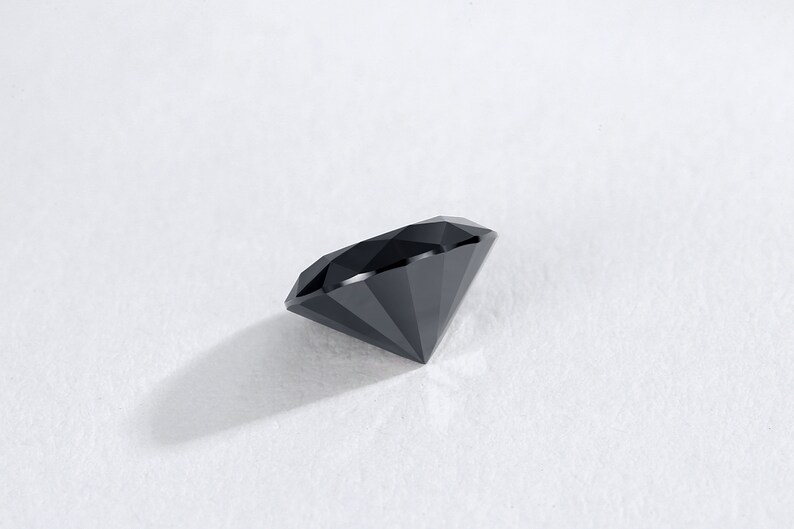 May include: A faceted, black gemstone with a brilliant cut. The diamond-shaped gem is displayed on a white surface, showcasing its sharp angles and reflective surfaces. The gemstone's dark color contrasts with the bright background.