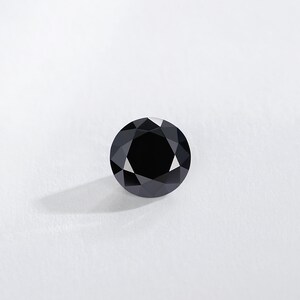 May include: A round, black gemstone with a faceted surface. The gem is set against a white background, with a shadow cast to the lower left. The gemstone is a deep, solid black color.