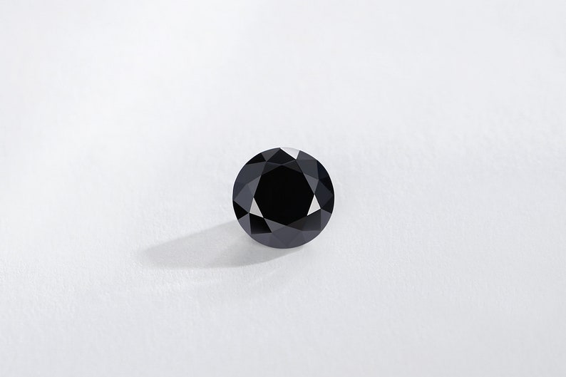May include: A round, black gemstone with a faceted surface. The gem is set against a white background, with a shadow cast to the lower left. The gemstone is a deep, solid black color.
