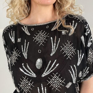 Può includere: Top nero con paillettes e perline argentate. Il top a maniche corte presenta uno scollo a barchetta ed è decorato con motivi geometrici e a forma di ventaglio. Le paillettes e le perline argentate creano un effetto scintillante sul tessuto nero.