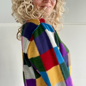 Peut inclure: Un pull patchwork coloré avec un col montant, présentant des motifs carrés dans des tons de bleu, violet, vert, jaune, rouge et gris. Le pull à manches longues arbore un design accrocheur et coloré.