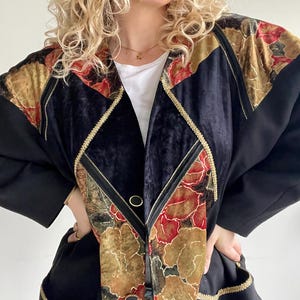 Op de afbeelding: Zwarte blazer met gouden bies en bloempatronen in rood, goud en groen. De blazer heeft een fluwelen textuur op de bovenborst en schouders, met twee gouden knopen en zakken.