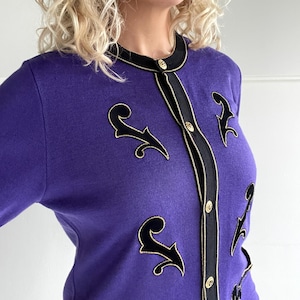 Op de afbeelding: Een paarse cardigan met zwarte bies en gouden knopen. De cardigan heeft een zwart fluwelen ontwerp aan de voorkant.