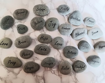 May include: A collection of painted stones with inspirational words like "Love", "Hope", "Joy", "Faith", "Peace", "Gratitude", "Family", "Courage", "Believe", "Trust", "Strength", "Happiness", "Dream", "Focus", "Friends", "Blessed", "Balance", "Inspire", "Pray", "Relax", "Thankful", "Imagine", "Success", "Brave", "Kindness", and "Laugh".