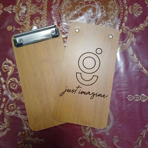 May include: Two wooden clipboards with a light brown wood grain finish. One clipboard has a metal clip at the top. The other has a logo and the words "just imagine" in a script font.