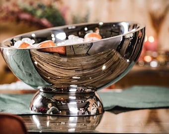 May include: A large, polished silver serving bowl with a wide, angled rim. The bowl sits on a pedestal base and reflects the surrounding environment. The bowl contains ice and orange-colored fruit. The bowl is on a wooden table.