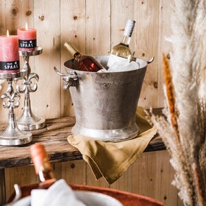 May include: A silver metal ice bucket with bull head handles, filled with ice and two bottles of wine. The bucket is sitting on a brown wooden surface with a gold cloth napkin. Two silver candle holders with candles are on the left side of the image.