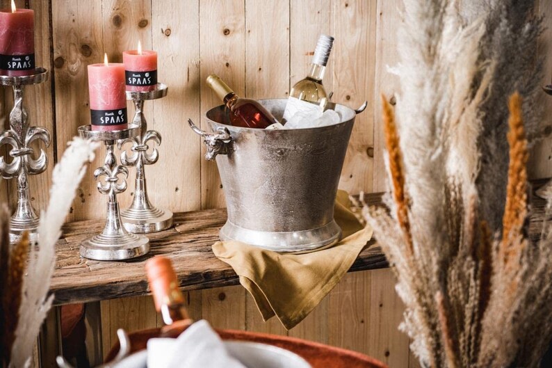 May include: A silver metal ice bucket with bull head handles, filled with ice and two bottles of wine. The bucket is sitting on a brown wooden surface with a gold cloth napkin. Two silver candle holders with candles are on the left side of the image.