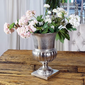 Peut inclure: Un vase argenté en forme d'urne rempli de fleurs roses et blanches et de feuilles vertes. Le vase est posé sur une base carrée et placé sur une table en bois. Les fleurs sont en pleine floraison.