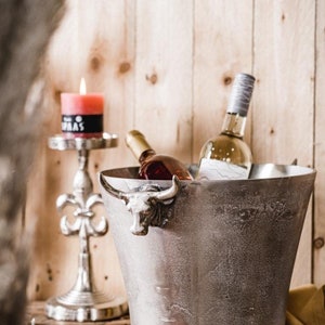 May include: A silver ice bucket with a bull head handle, holding two bottles of wine. The ice bucket is sitting on a brown wooden surface with a gold candle holder and a lit candle in the background.