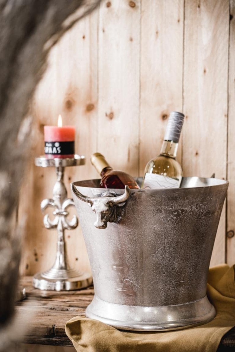 May include: A silver ice bucket with a bull head handle, holding two bottles of wine. The ice bucket is sitting on a brown wooden surface with a gold candle holder and a lit candle in the background.