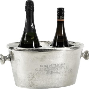 May include: Silver metal champagne bucket with two bottles of champagne inside. The bucket has a handle on each side and the text "Cuv&eacute;e de Prestige Champagne De Laurance" is engraved on the front.