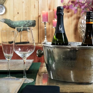 May include: A silver metal ice bucket with two bottles of wine inside. The ice bucket has the words "Coffre de Prestige" engraved on the side. The bottles are in a dark green and brown glass.