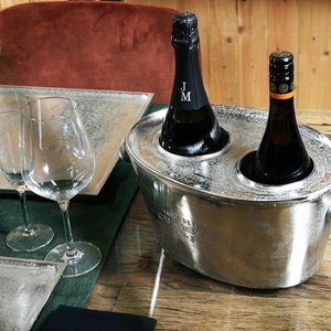 May include: A silver metal wine cooler with two bottles of wine inside. The cooler has a handle on the side and a spout on the top. The bottles are both dark green glass with labels. The cooler is sitting on a wooden table.