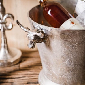 May include: Silver ice bucket with a bull head handle, filled with a bottle of rose wine. The bucket is sitting on a wooden surface with a brown cloth napkin.
