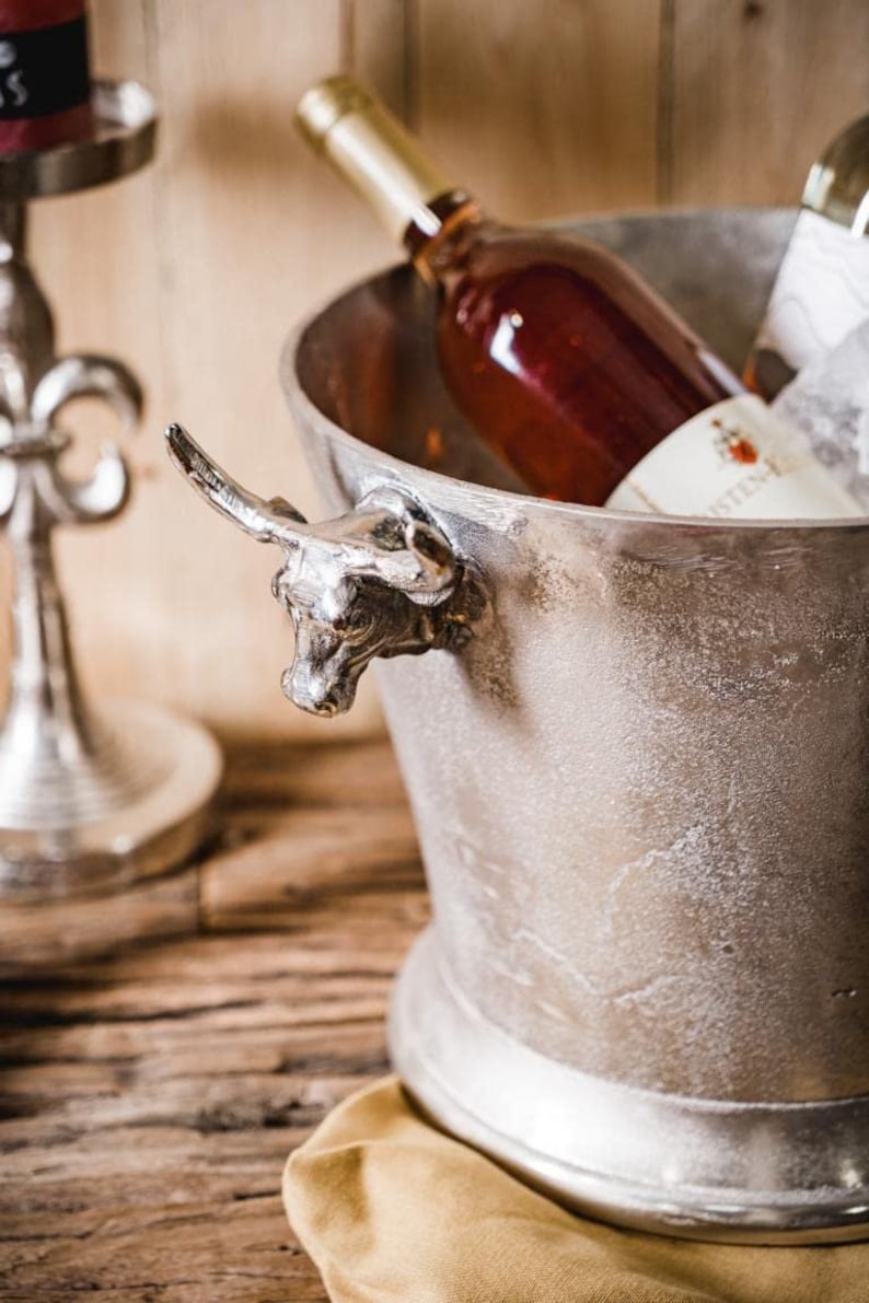 May include: Silver ice bucket with a bull head handle, filled with a bottle of rose wine. The bucket is sitting on a wooden surface with a brown cloth napkin.
